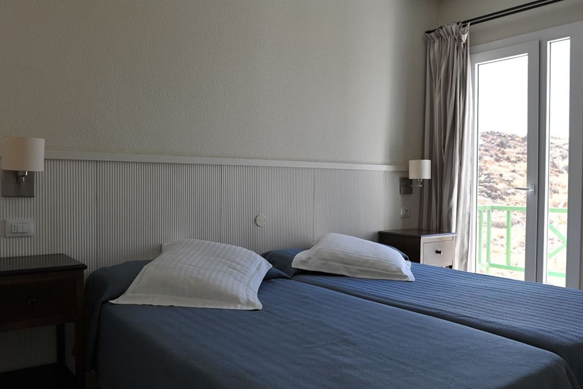 Habitación de hotel con dos camas, una mesita de noche, una lámpara y una ventana con cortinas.
