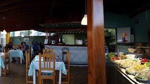 Un restaurante con mesas y sillas de madera, un mostrador de buffet con diversos platos, y una zona de cocina al fondo.
