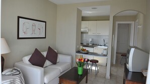 Una sala de estar con un sofá, una mesa de centro con un jarrón de flores, una zona de cocina con armarios y un refrigerador, y un televisor sobre un mueble.