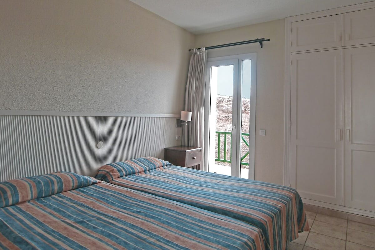 Una habitación de hotel con cama, mesita de noche, lámpara, puerta corrediza de vidrio a un balcón y un armario.