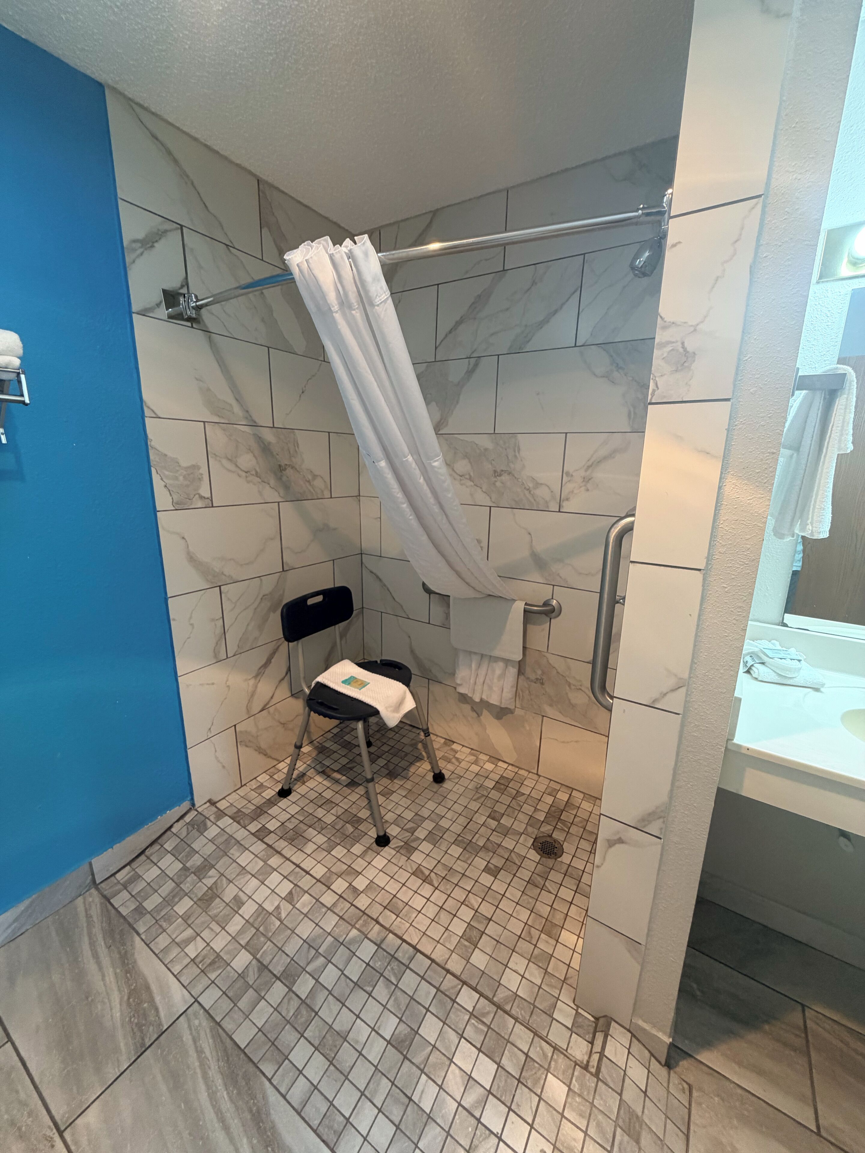 A shower stall with a white shower curtain, a small stool, a handrail, and a tiled floor.