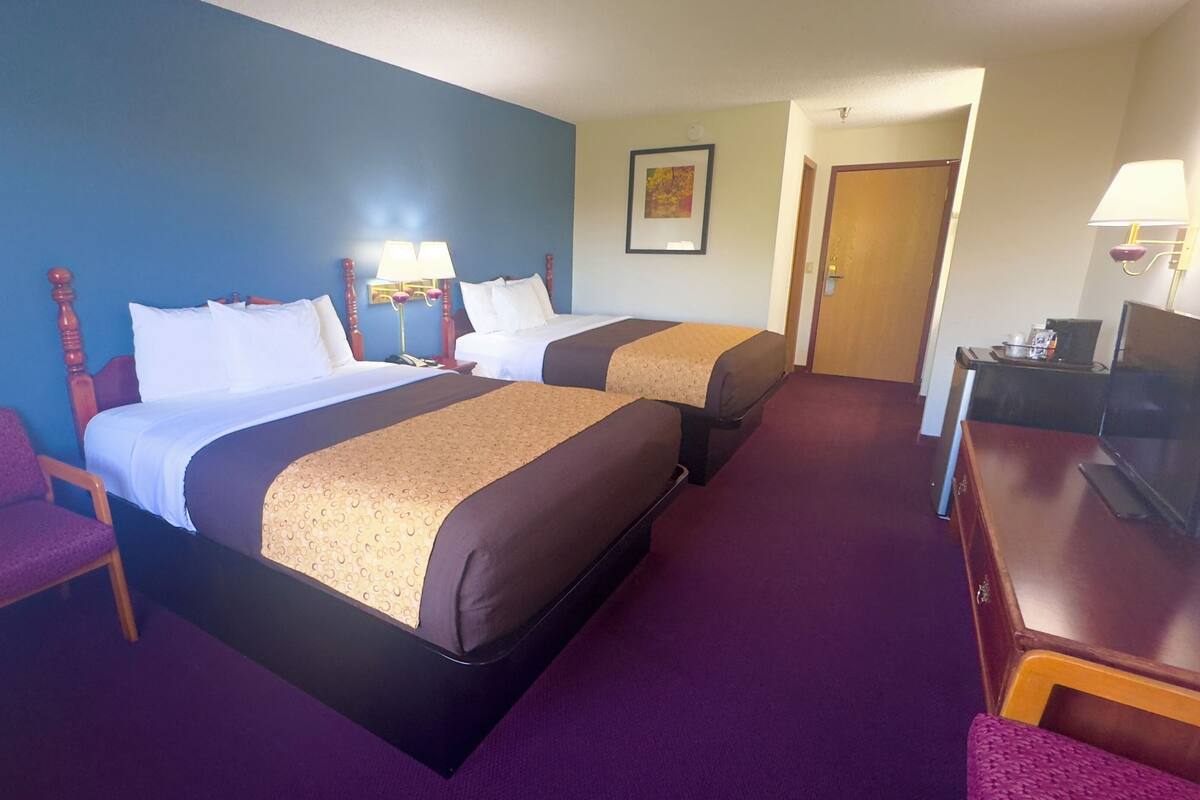 A hotel room with two beds, a desk with a computer, a chair, and a door leading to another room.
