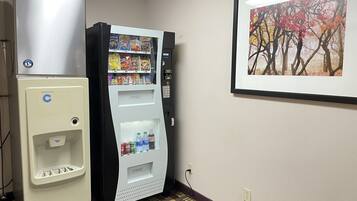A vending machine with snacks and drinks, a refrigerator, and a framed picture on the wall.