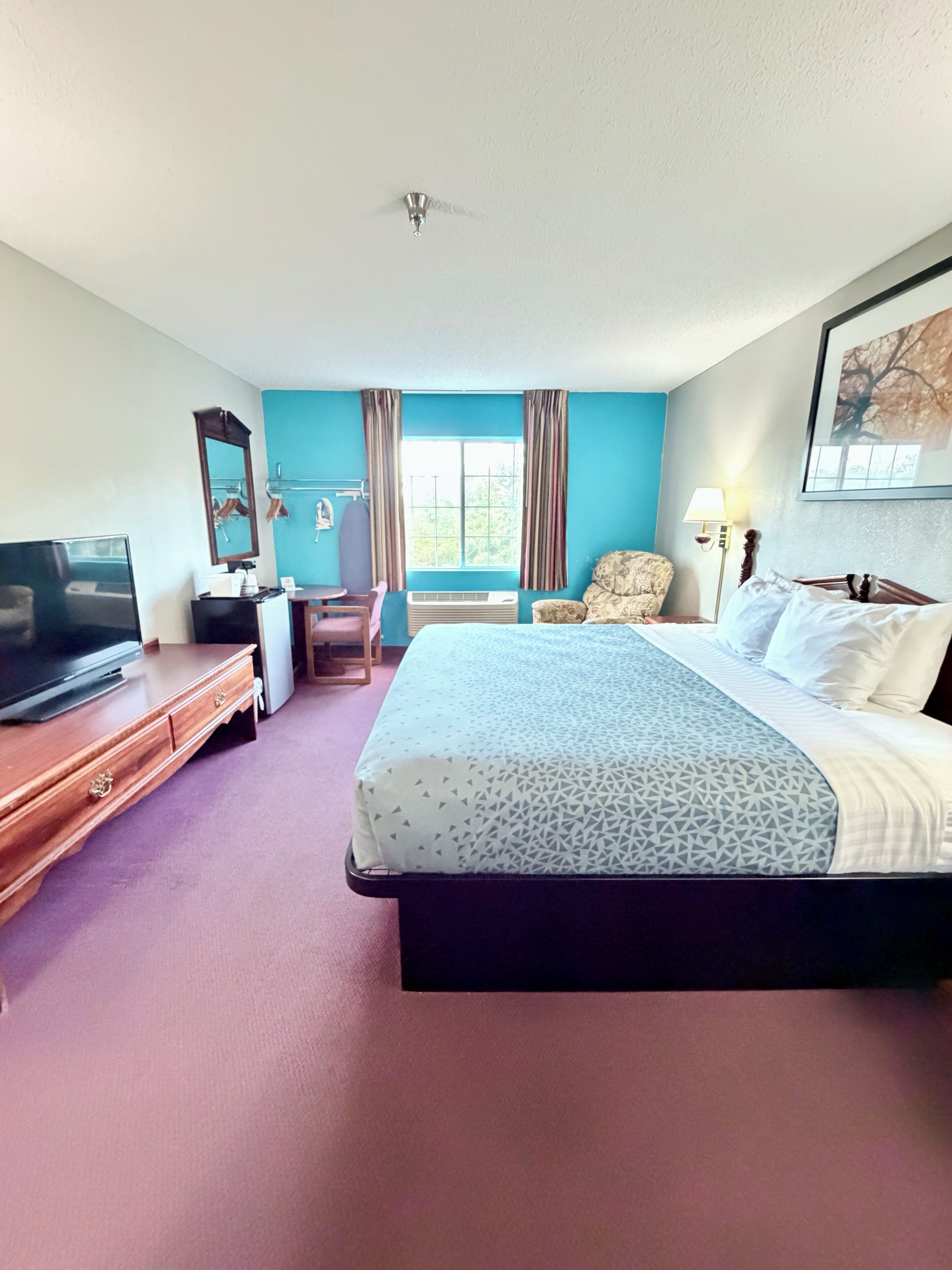 A hotel room with a bed, a TV, a desk, a chair, a lamp, and a window with curtains.