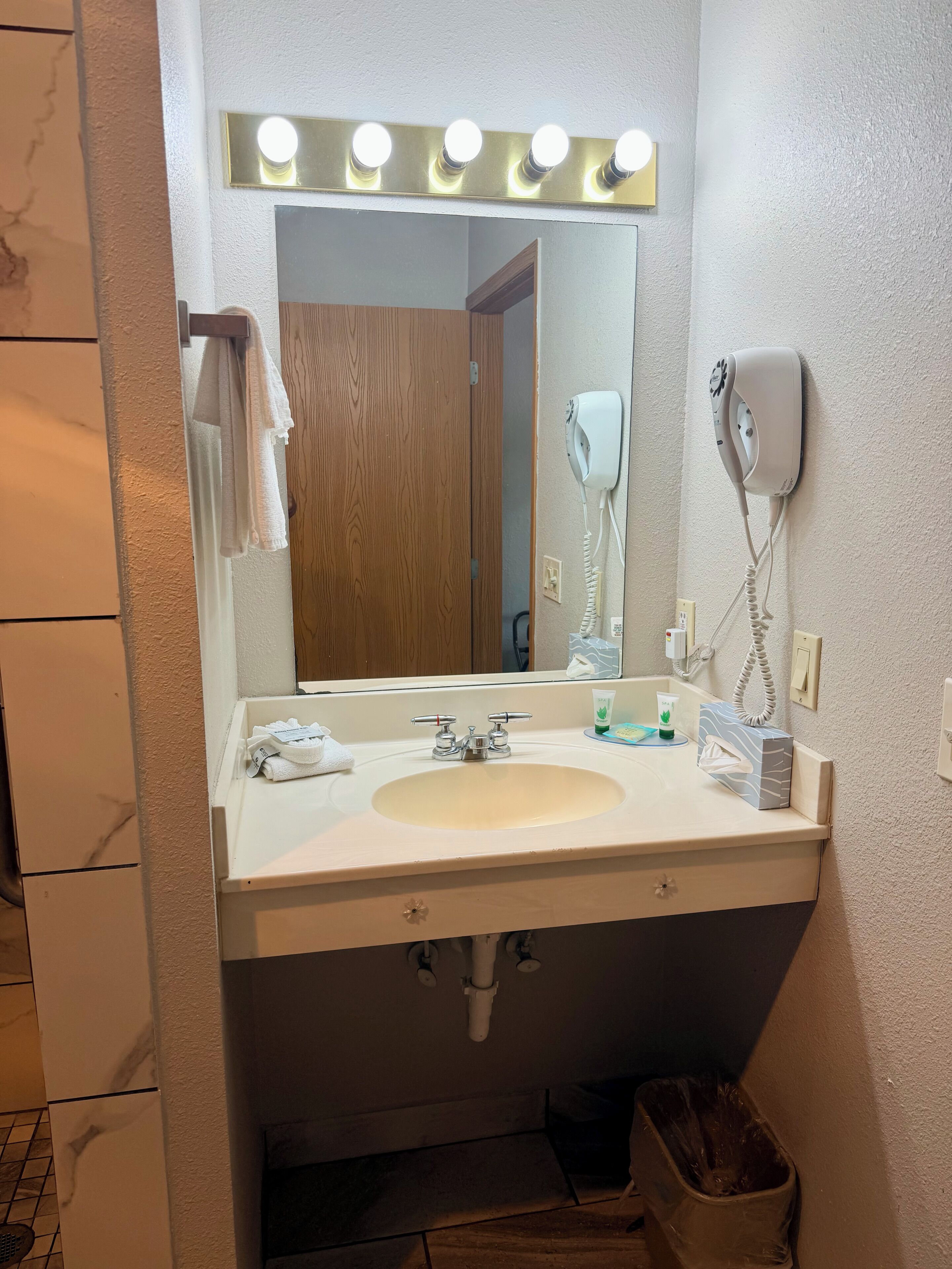 A bathroom with a sink, mirror, vanity lights, a hairdryer, and a trash bin.