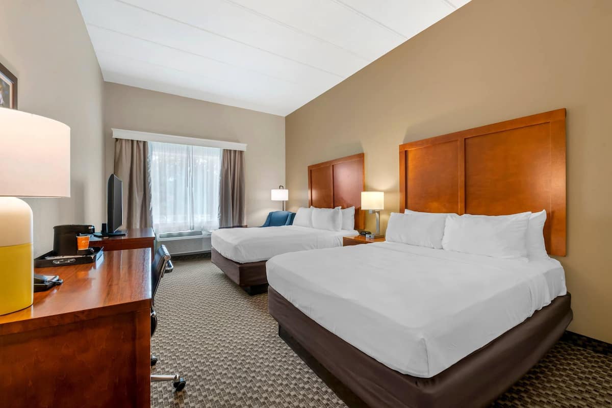 Ein Hotelzimmer mit zwei Betten, einem Schreibtisch mit Lampe, einem Fernseher und einem Fenster mit Vorhängen.