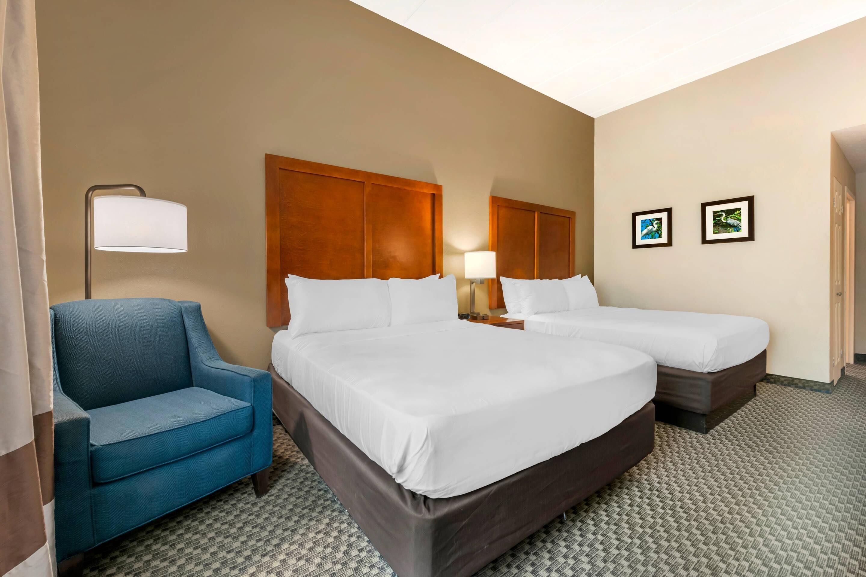 A hotel room with two beds, a blue armchair, a lamp, and two framed pictures on the wall.