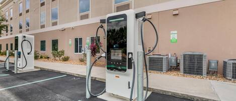 Un área de estacionamiento con estaciones de carga para vehículos eléctricos, un edificio con amplias ventanas y unidades de aire acondicionado al aire libre.