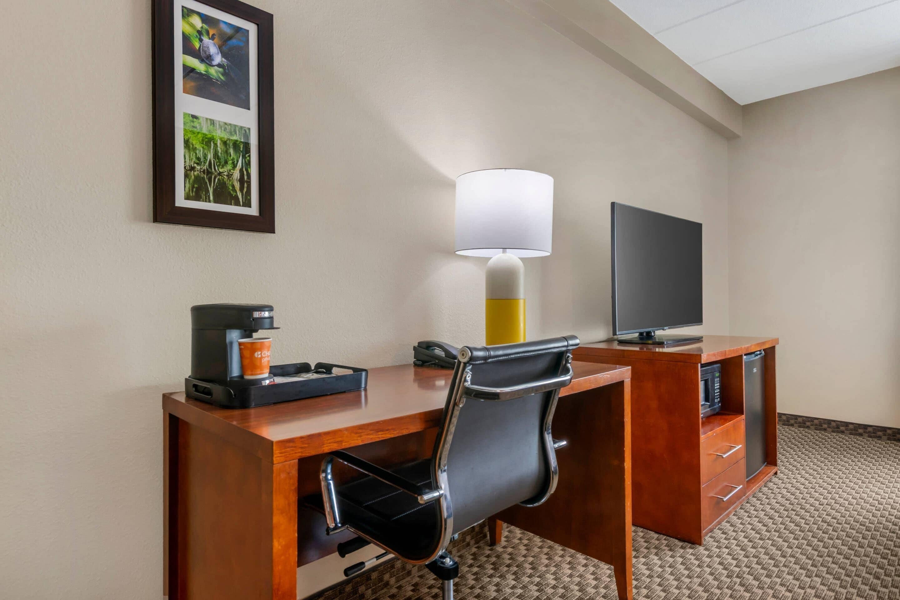 A hotel room with a wooden desk, a black office chair, a coffee maker, a lamp, a flat-screen TV, and a framed picture on the wall.