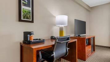 A hotel room with a wooden desk, a black office chair, a coffee maker, a lamp, a flat-screen TV, and a framed picture on the wall.