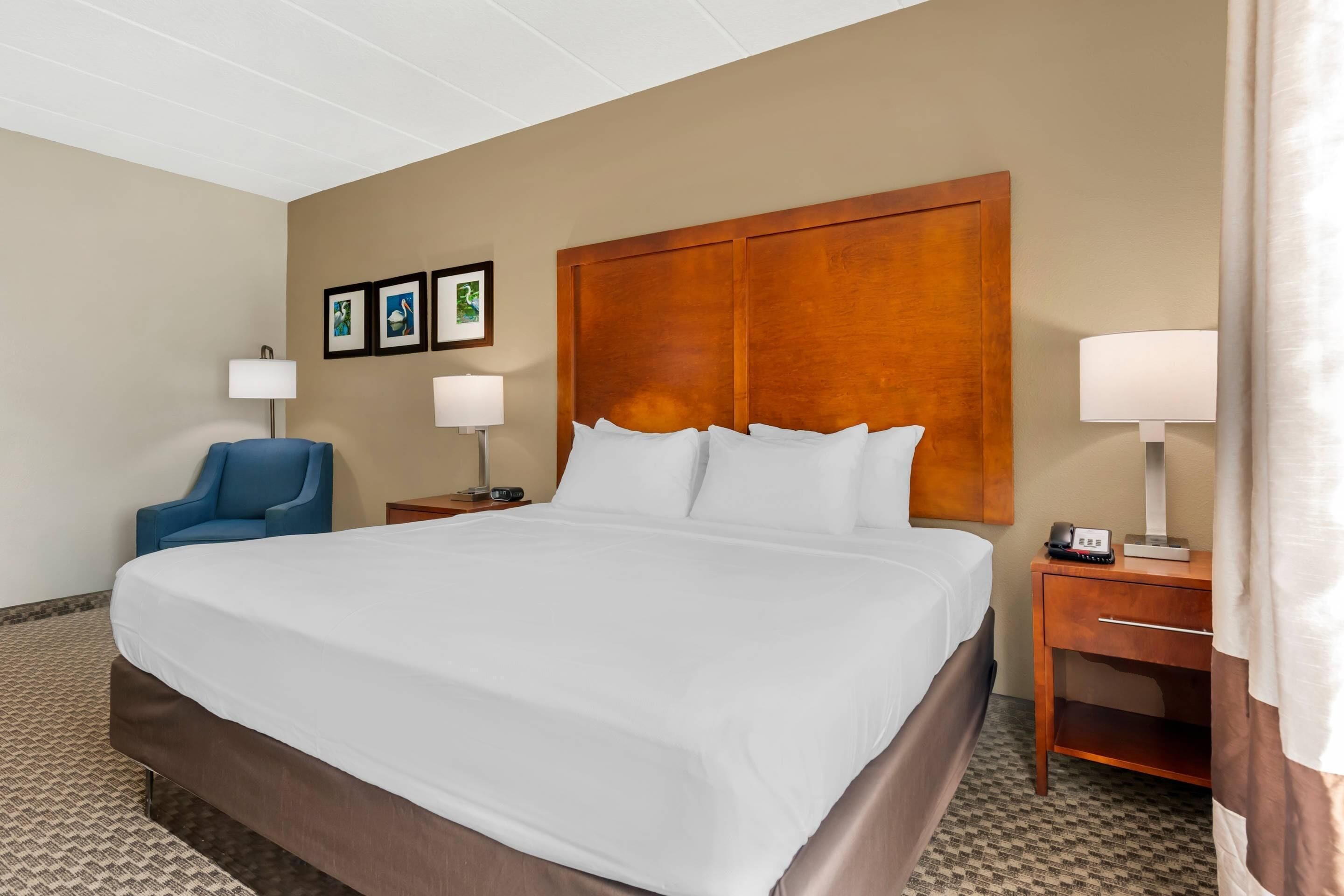Quarto de hotel com uma cama grande, duas mesas de cabeceira com abajures, uma cadeira azul e quadros nas paredes.