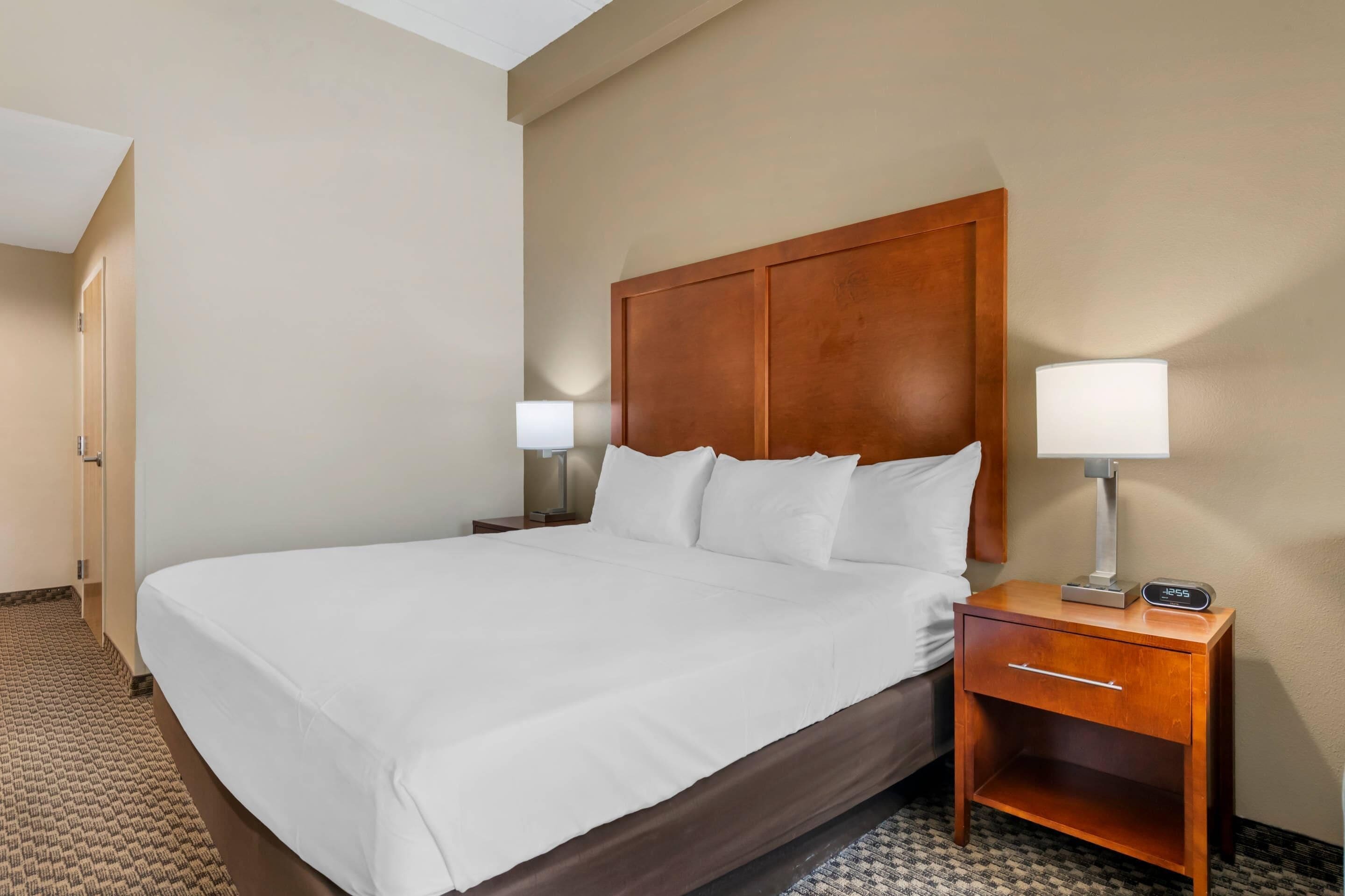 A hotel room with a large bed, a wooden headboard, a bedside table with a lamp, and a nightstand with a clock.