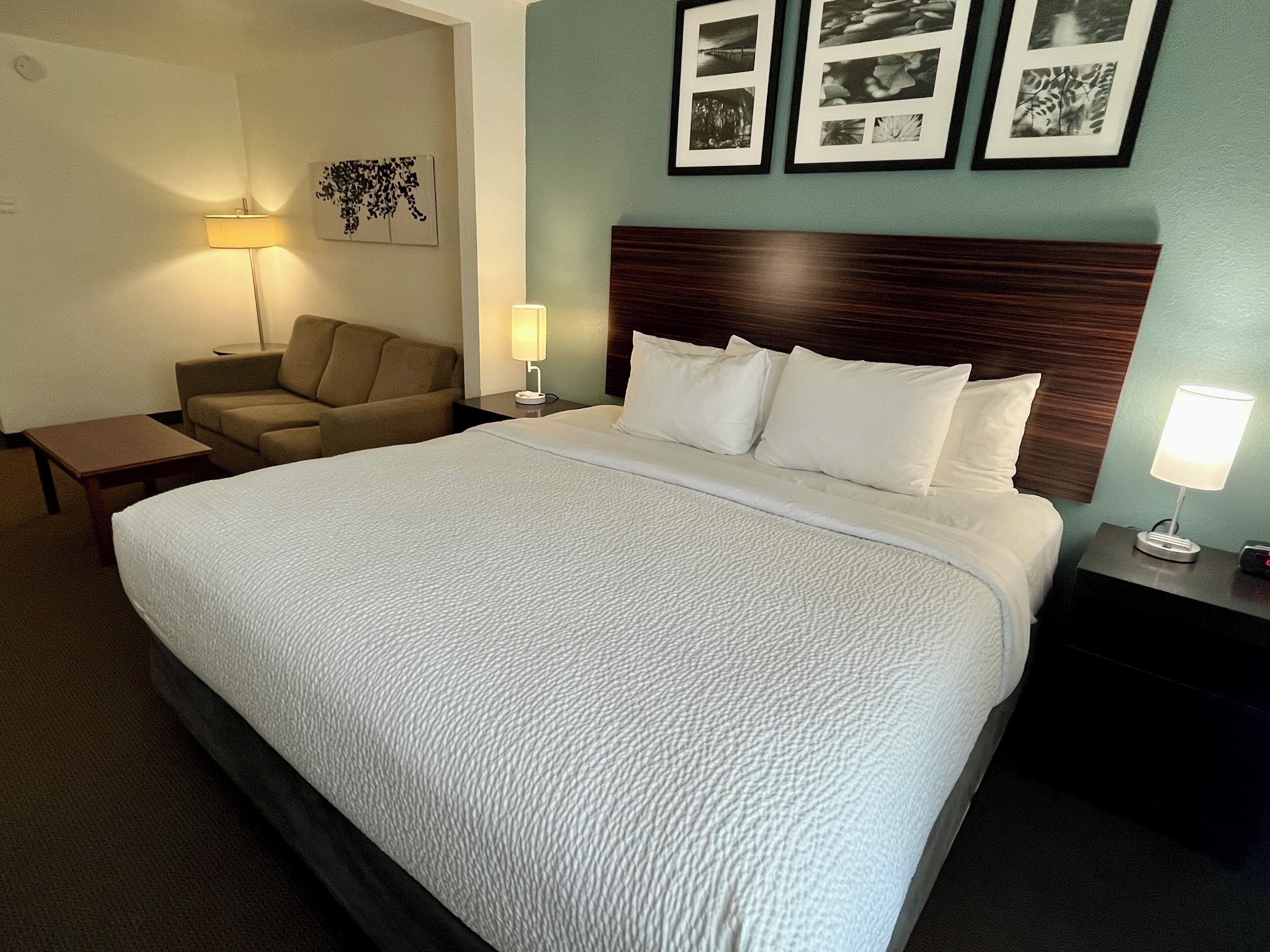 A hotel room with a large bed, a nightstand with a lamp, a chair, and a wall with framed pictures.