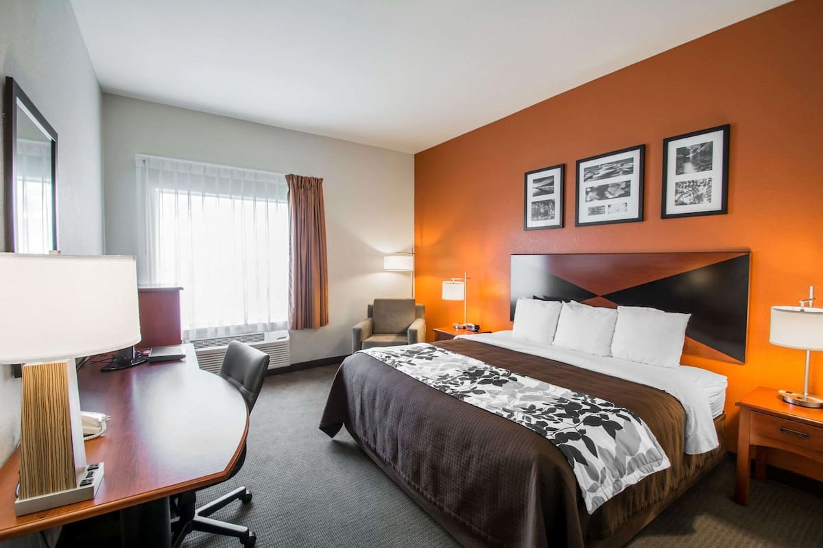 A hotel room with a large bed, a desk with a lamp, a chair, and a window with curtains.