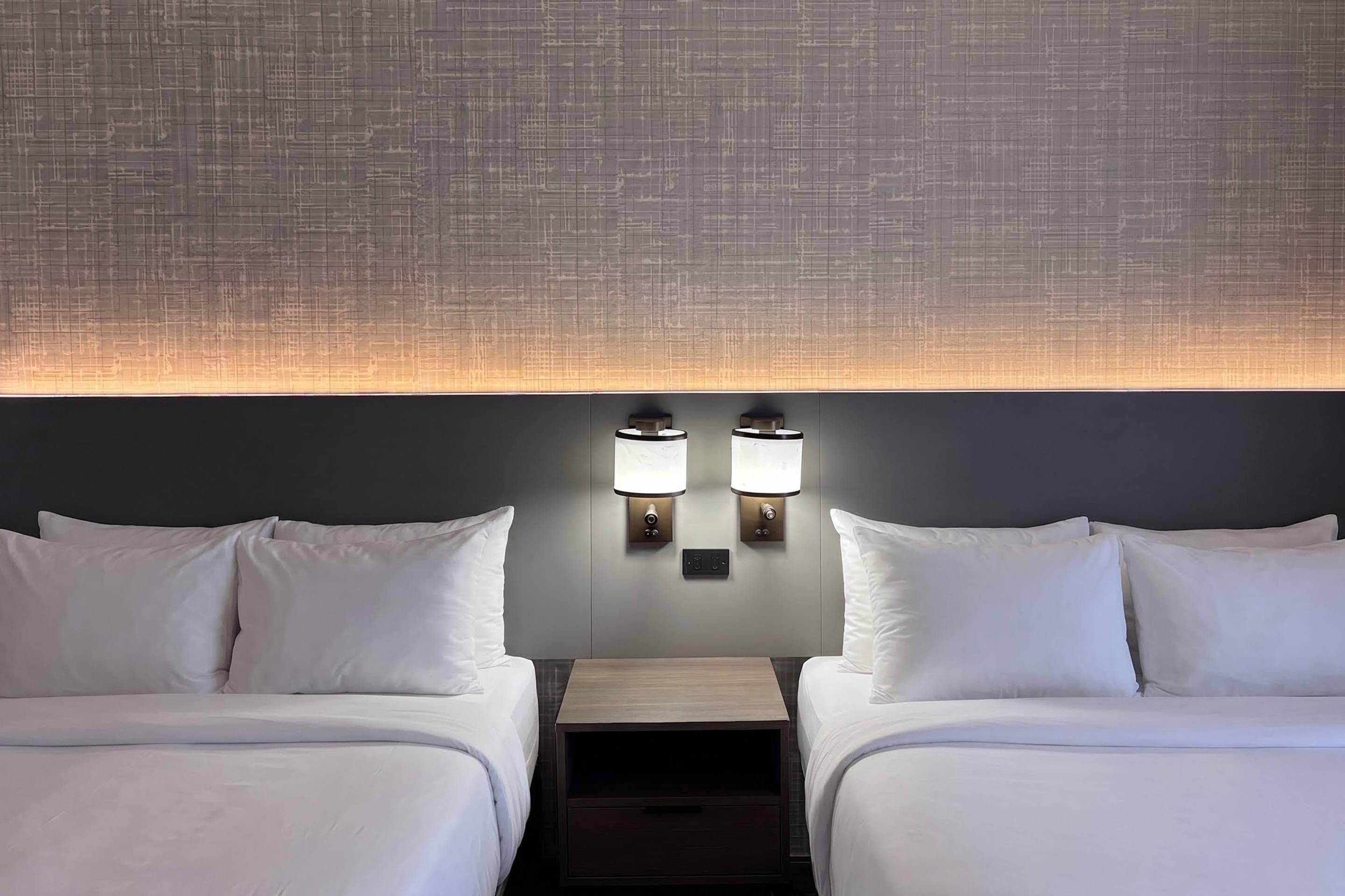 A hotel room with two beds, a nightstand, and wall-mounted lamps.