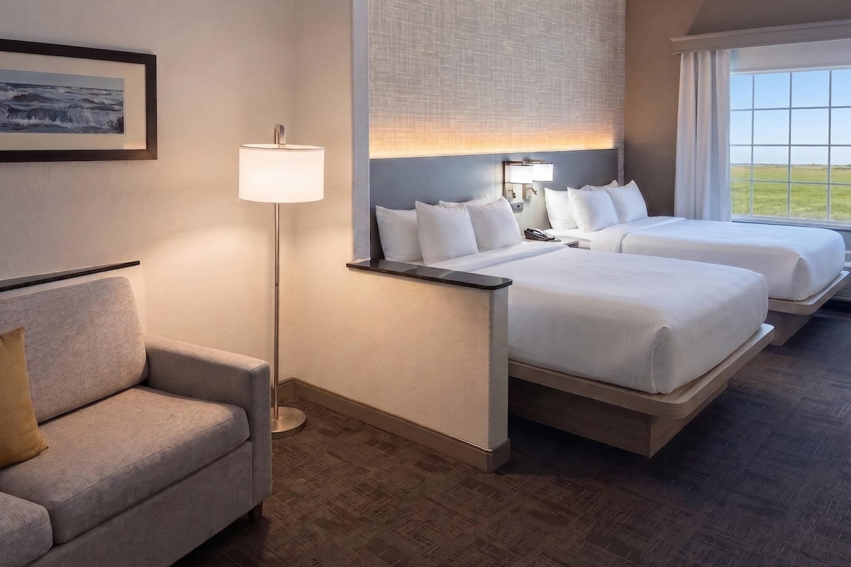A hotel room with two beds, a sofa, a floor lamp, and a window with a view.