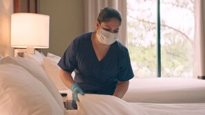 A person in a dark scrub top and gloves making a bed in a hotel room with white bedding and a lamp on the side table. - Comfort Suites Galveston Cruise Port Area – Beachside (Galveston)