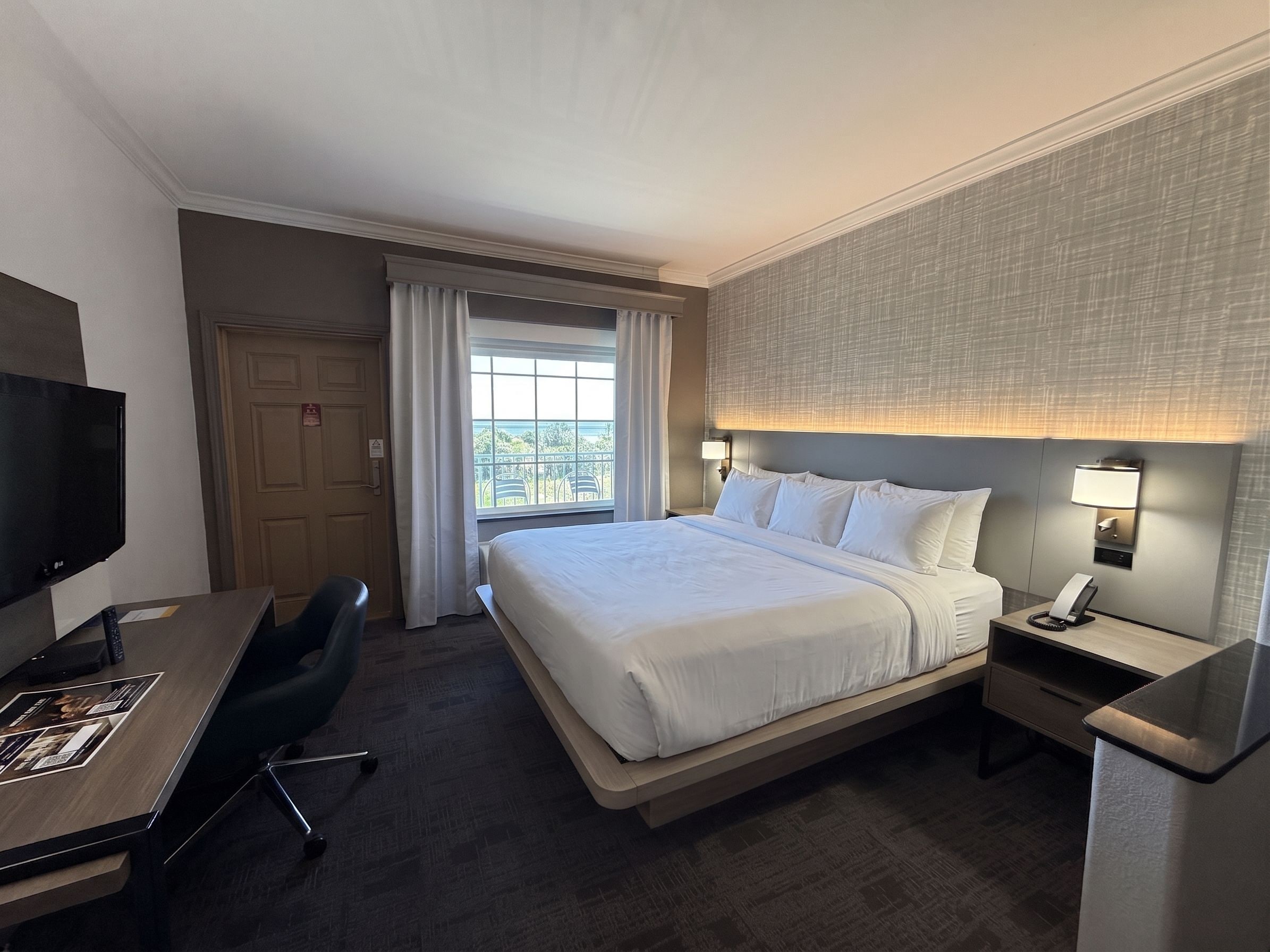 Quarto de hotel com cama, escrivaninha, cadeira, televisão e vista para o exterior.