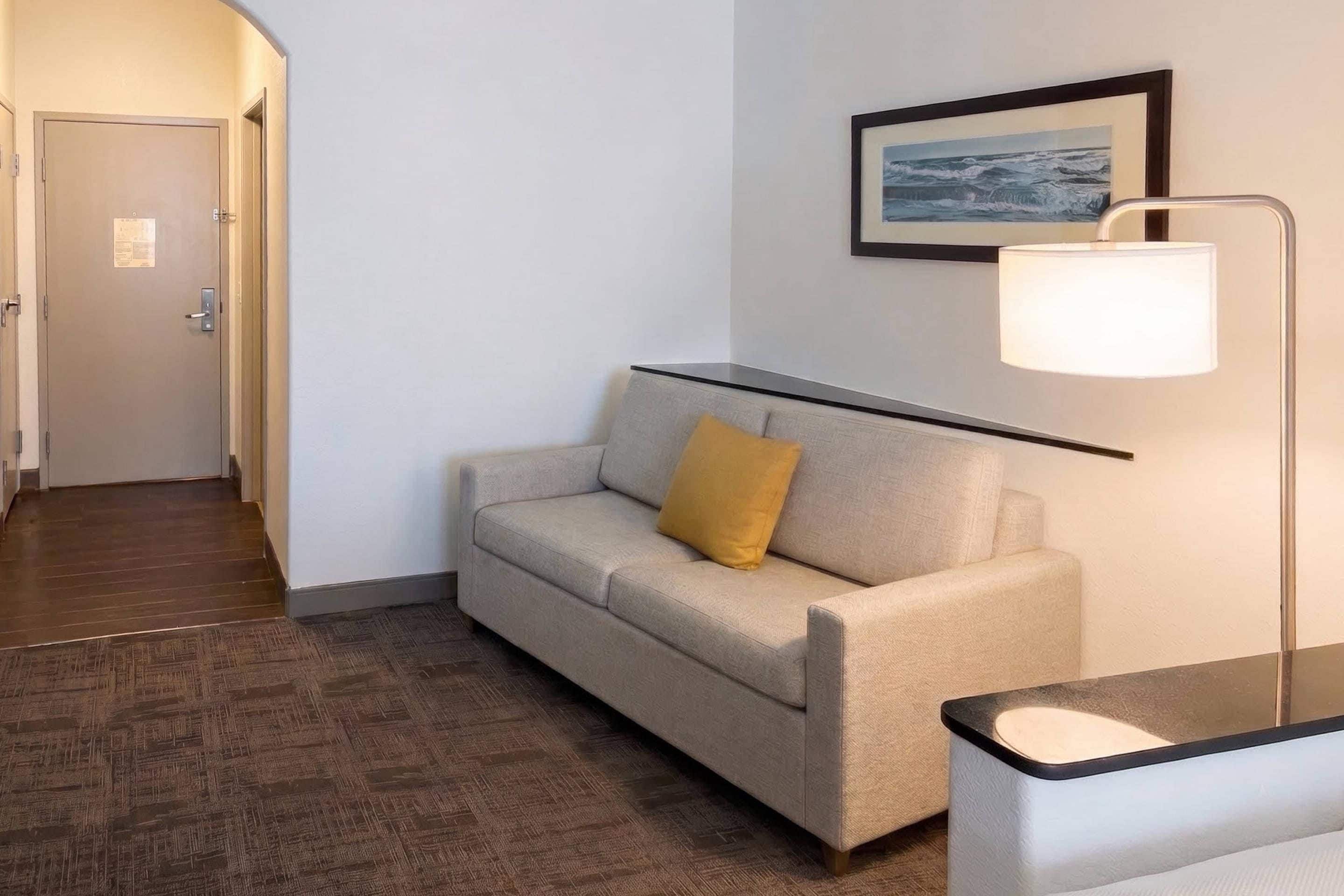 Quarto de hotel com sofá, luminária, quadro na parede e piso de madeira.