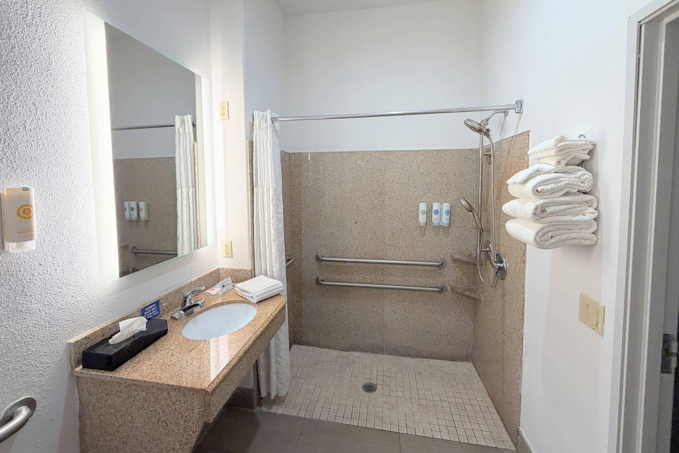 A bathroom with a sink, mirror, towel rack, shower, and handrails.