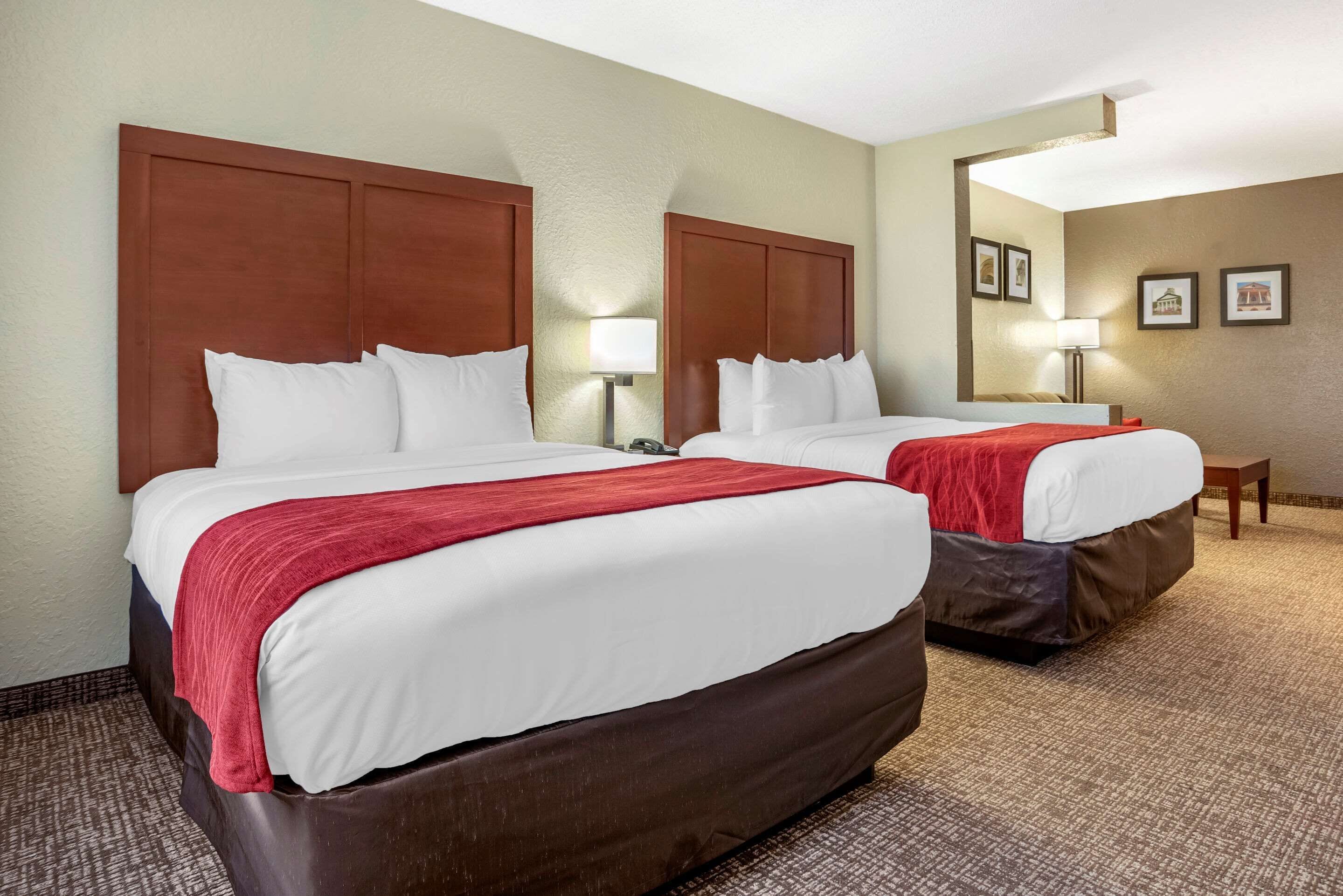 A hotel room with two beds, each with a wooden headboard, white bedding, and a red throw blanket. There is a nightstand with a lamp, a small table, and framed pictures on the wall.
