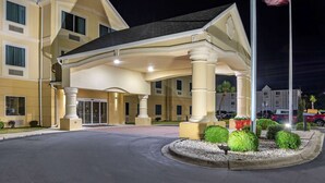 Un hotel con entrada cubierta, un asta con la bandera estadounidense y una zona ajardinada con arbustos.