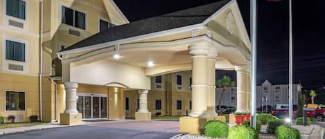 A hotel with a covered entrance, a flagpole with an American flag, and a landscaped area with shrubs.