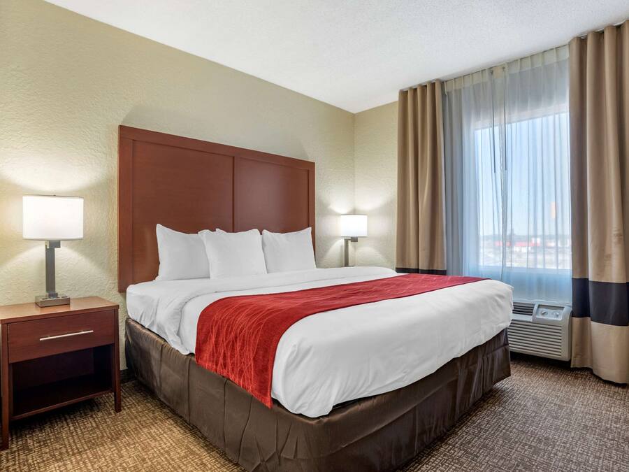 A hotel room with a large bed, two bedside tables with lamps, a window with curtains, and an air conditioning unit.
