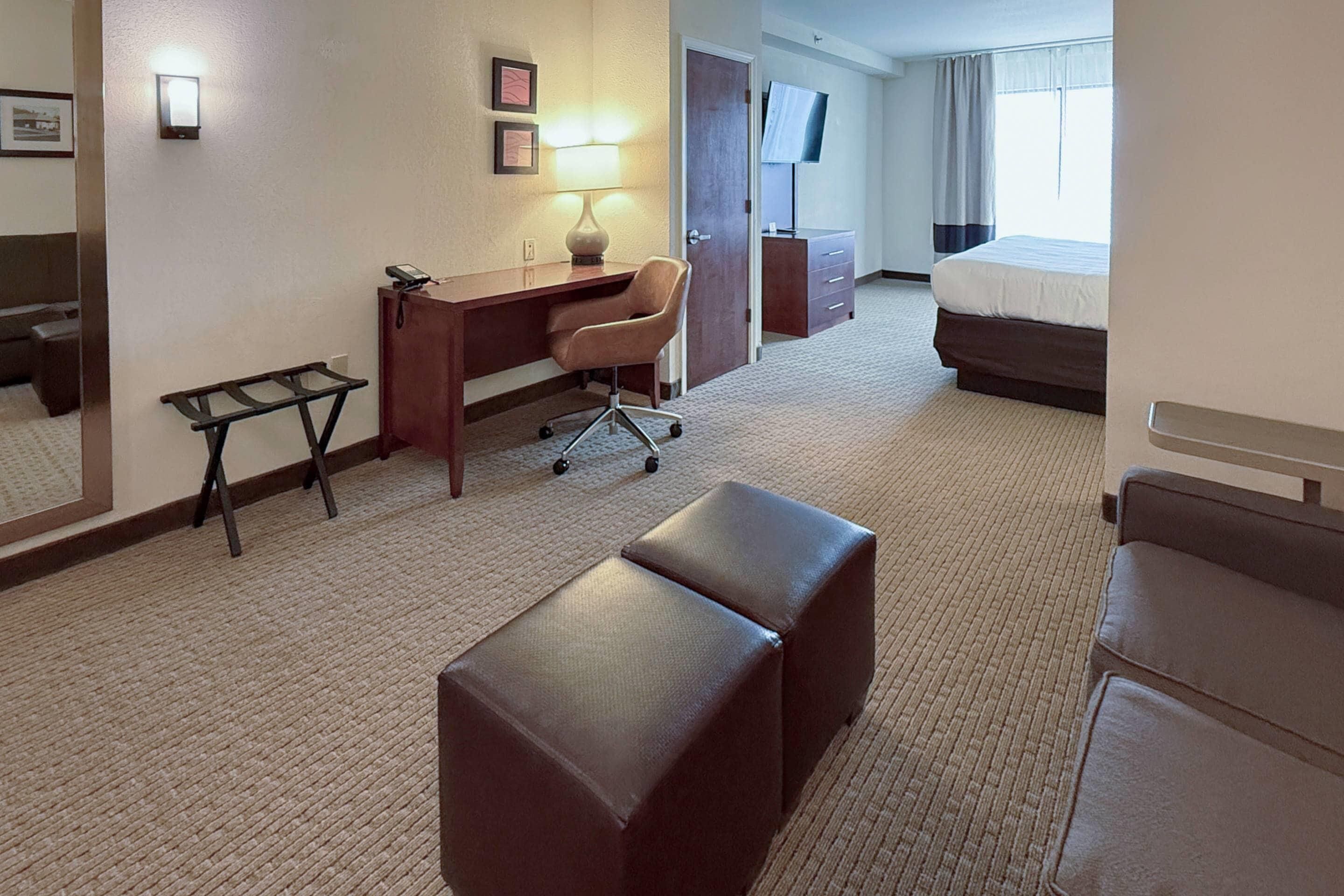 A hotel room with a desk, chair, ottoman, bed, TV, and a door leading to another room.