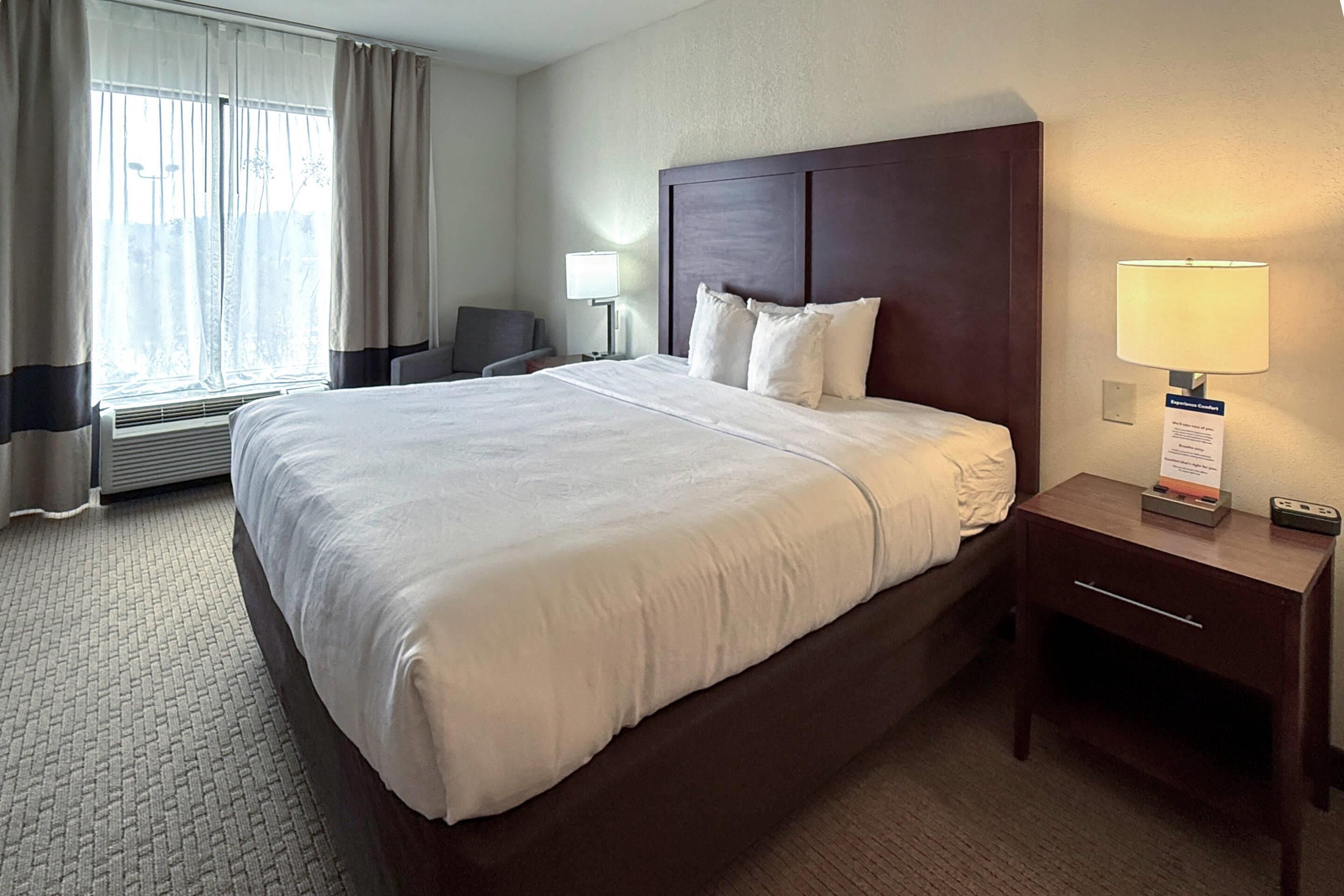 A hotel room with a large bed, a bedside table with a lamp, a chair, and a window with curtains.