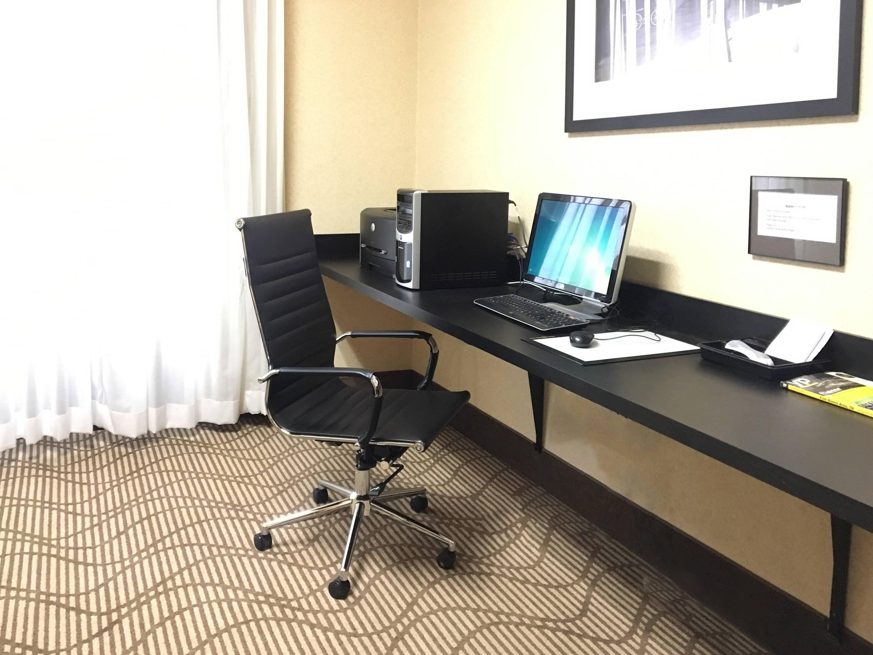 A hotel office with a desk, chair, computer, and framed picture on the wall.