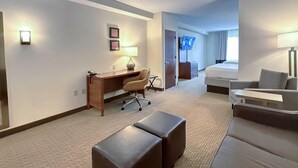 A hotel room with a desk, office chair, two ottomans, a sofa, a TV, and a bed.