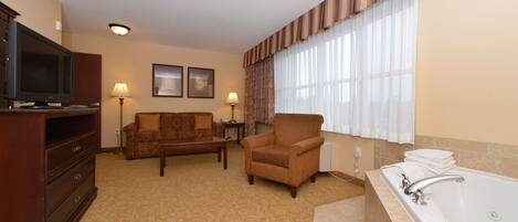 Habitación de hotel con televisión de pantalla plana, sofá, sillón, pequeña mesa, lámpara, ventana con cortinas y bañera.