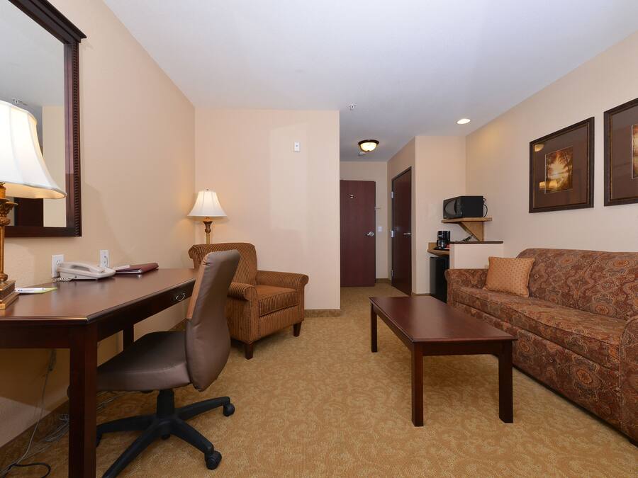 A hotel room with a desk, office chair, sofa, and a small table. There is a lamp on the desk, a telephone, and a picture frame on the wall.