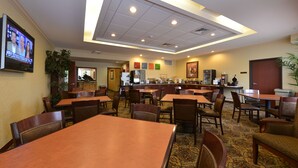 Un'area ristorazione in hotel con diversi tavoli e sedie, una televisione appesa al muro e una cucina sullo sfondo.