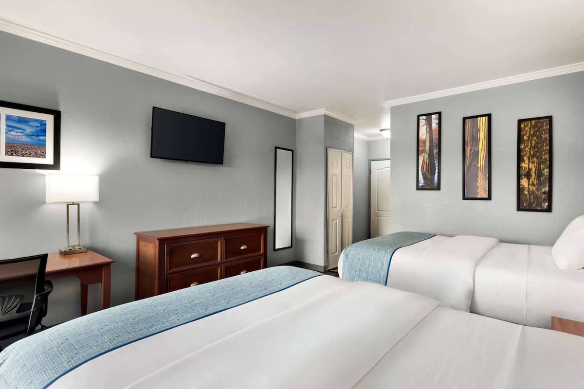 A hotel room with two beds, a desk, a chair, a dresser, a TV, and framed artwork on the wall.