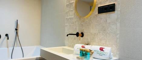 A bathroom with a white sink, a round mirror with a gold frame, and a towel dispenser with a hotel logo.