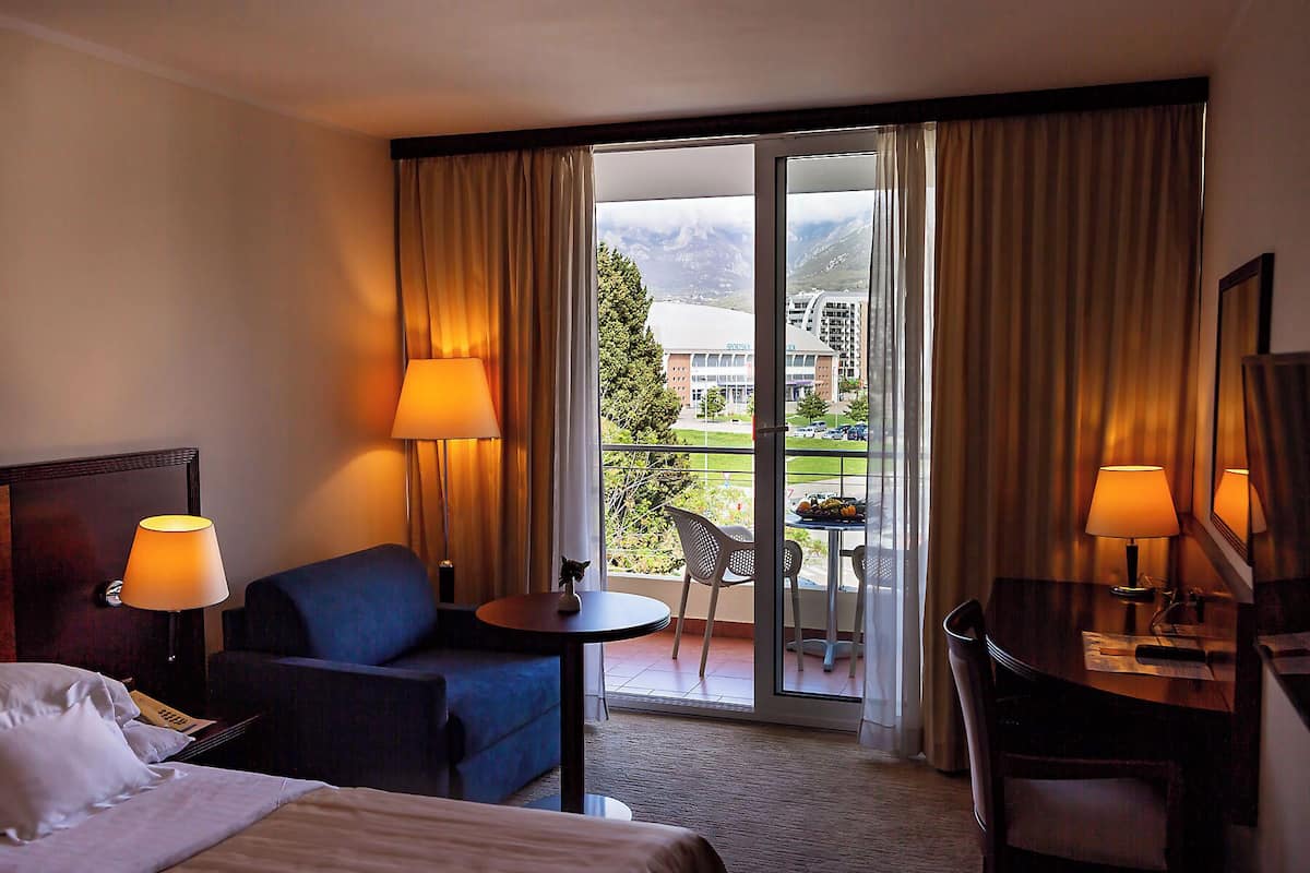 A hotel room with a bed, a blue armchair, a small table with a lamp, a dining table with chairs, and a view of a building and mountains through the window.