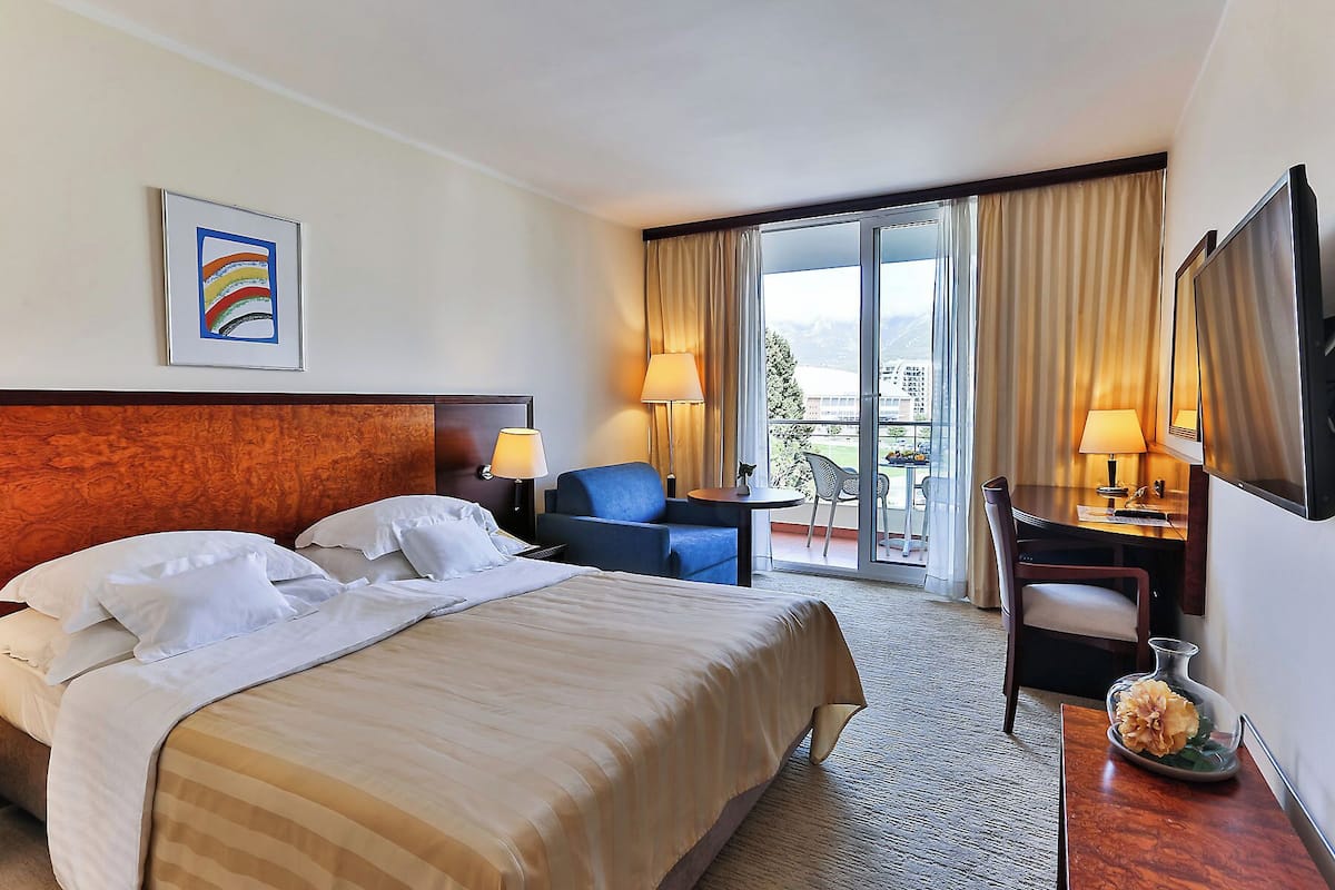 A hotel room with a bed, a desk, a chair, a TV, and a balcony with curtains.