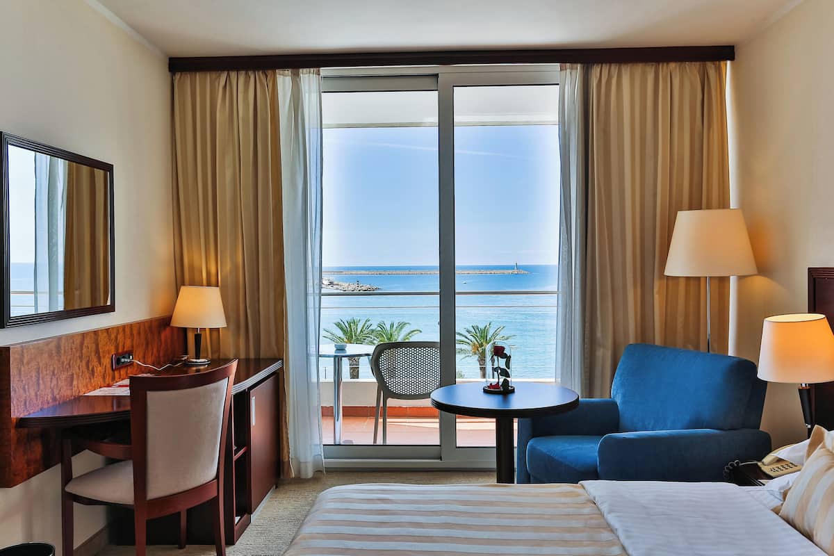 A hotel room with a bed, a desk, a chair, a blue sofa, a small table, and a view of the sea.