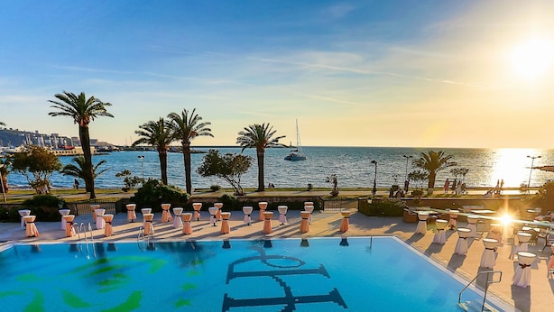 A pool with a view of the sea and boats, surrounded by palm trees and outdoor seating. - Hotel Princess (Bar)