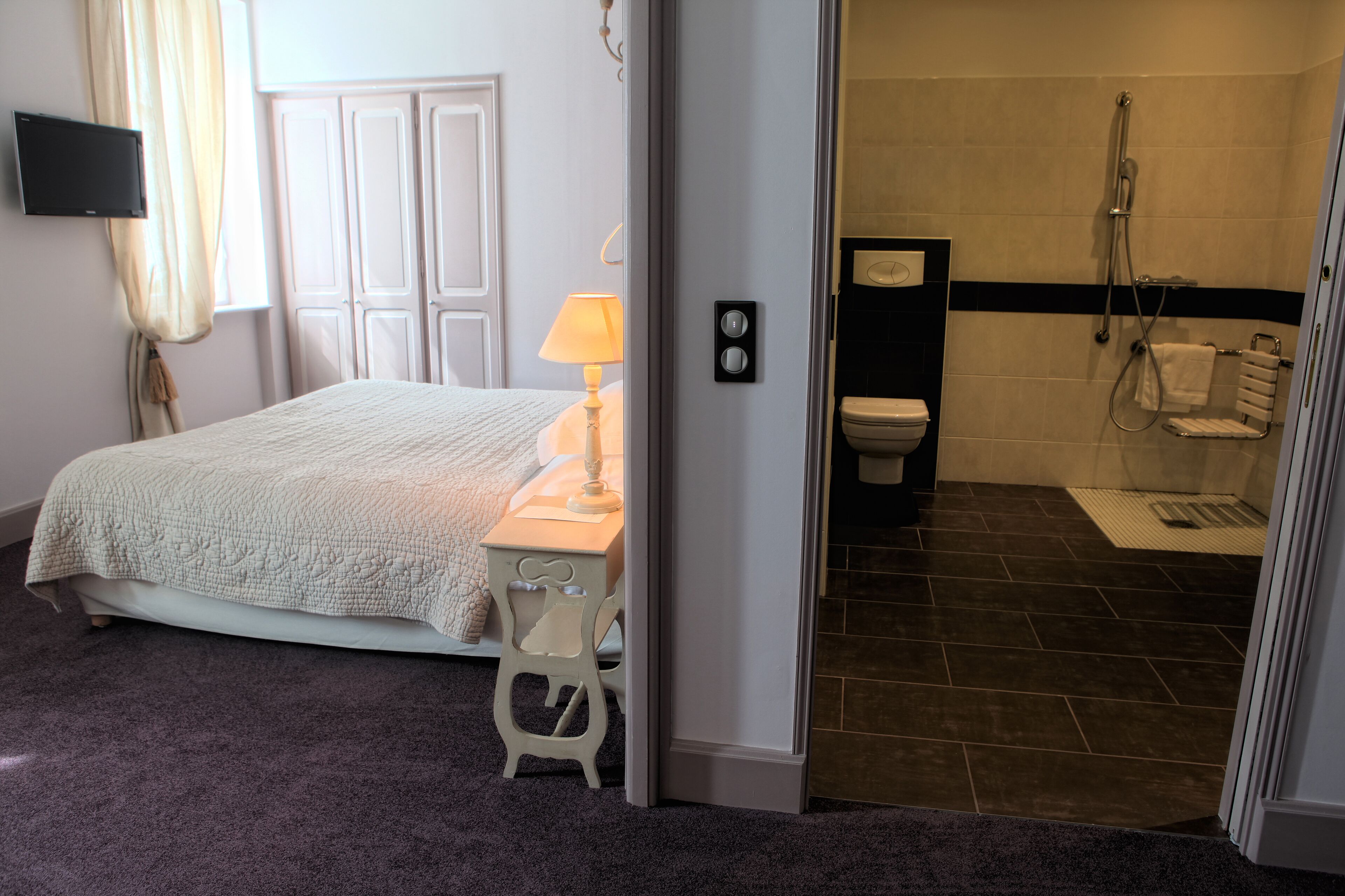 Een slaapkamer met een bed, nachtkastje, lamp en een deur naar een badkamer met douche en toilet.