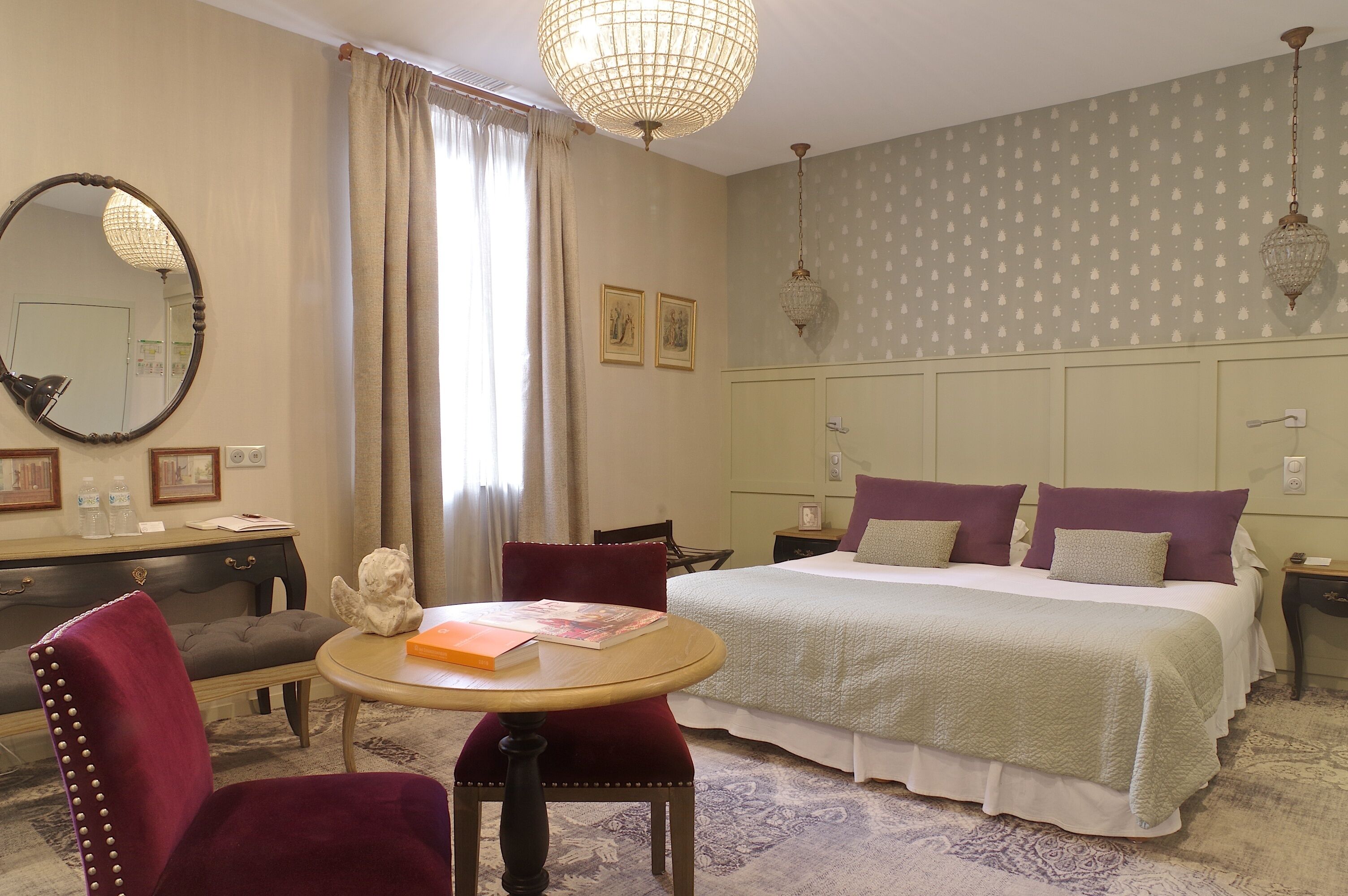 A hotel room with a bed, a round table, a chair, a desk, a mirror, and a chandelier.