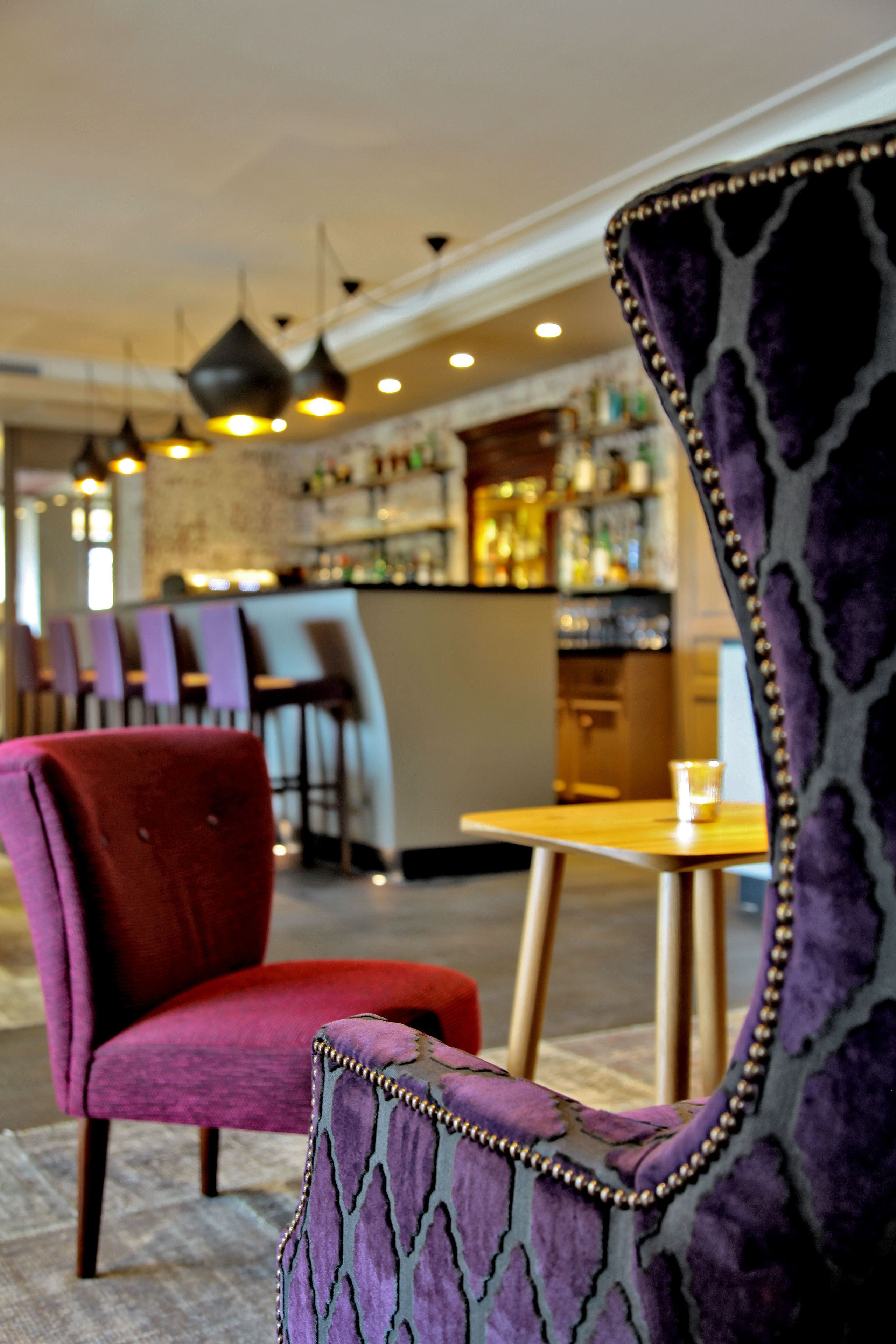 Een restaurantinterieur met een bar, rode en paarse stoelen met bekleding en hanglampen.