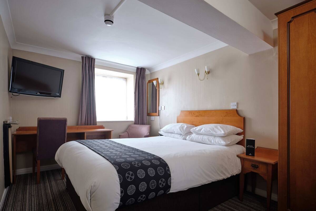 Quarto de hotel com cama, escrivaninha, cadeira, televisão e guarda-roupa.