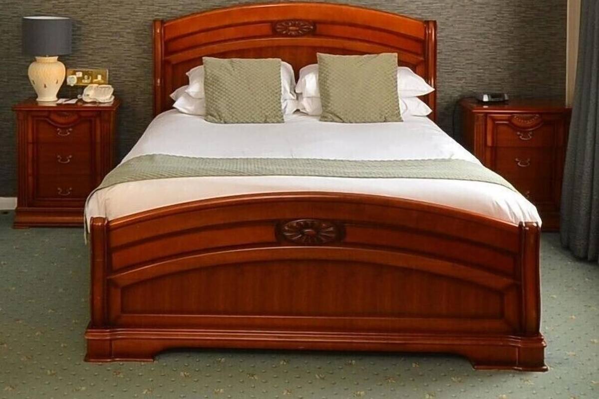 Uma cama de madeira com dossel, um painel de cabeceira estofado em verde, duas mesinhas de cabeceira e uma parede com textura.
