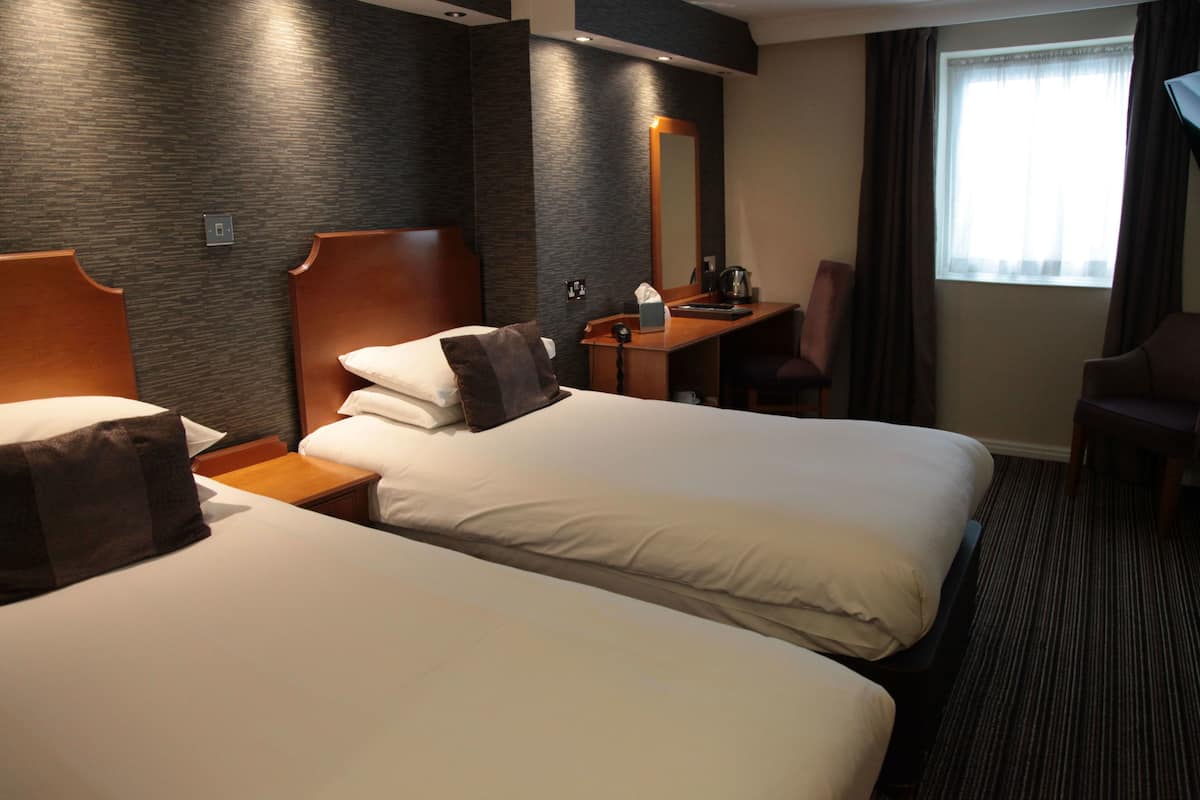 Quarto de hotel com duas camas, uma escrivaninha, uma cadeira e um espelho grande.
