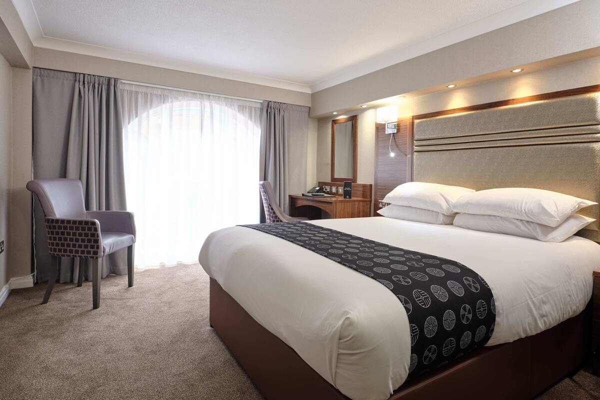 Quarto de hotel com uma cama grande, uma cadeira, uma escrivaninha com telefone e um espelho.