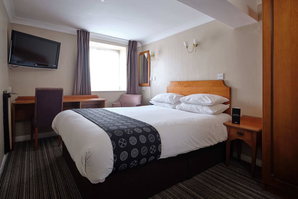 Quarto de hotel com cama, escrivaninha, cadeira, televisão e janela com cortinas.