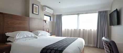 A hotel room with a large bed, a wooden headboard, a bedside table, a flat-screen TV mounted on the wall, and a window with curtains.